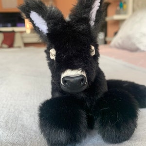 May include: A plush black donkey toy with large, fluffy ears with white inner ear details. The donkey has a white muzzle and a black nose. The toy is lying down on a soft, light-colored surface.