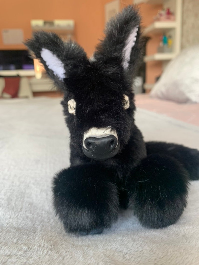 May include: A plush black donkey toy with large, fluffy ears with white inner ear details. The donkey has a white muzzle and a black nose. The toy is lying down on a soft, light-colored surface.