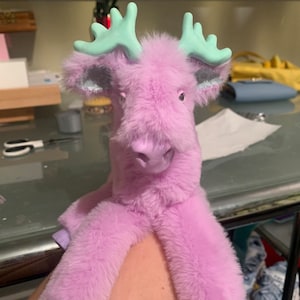 May include: A plush, lavender-colored moose toy with mint green antlers. The toy has a soft, furry texture and a friendly expression. The moose's hooves are a lighter shade of purple.