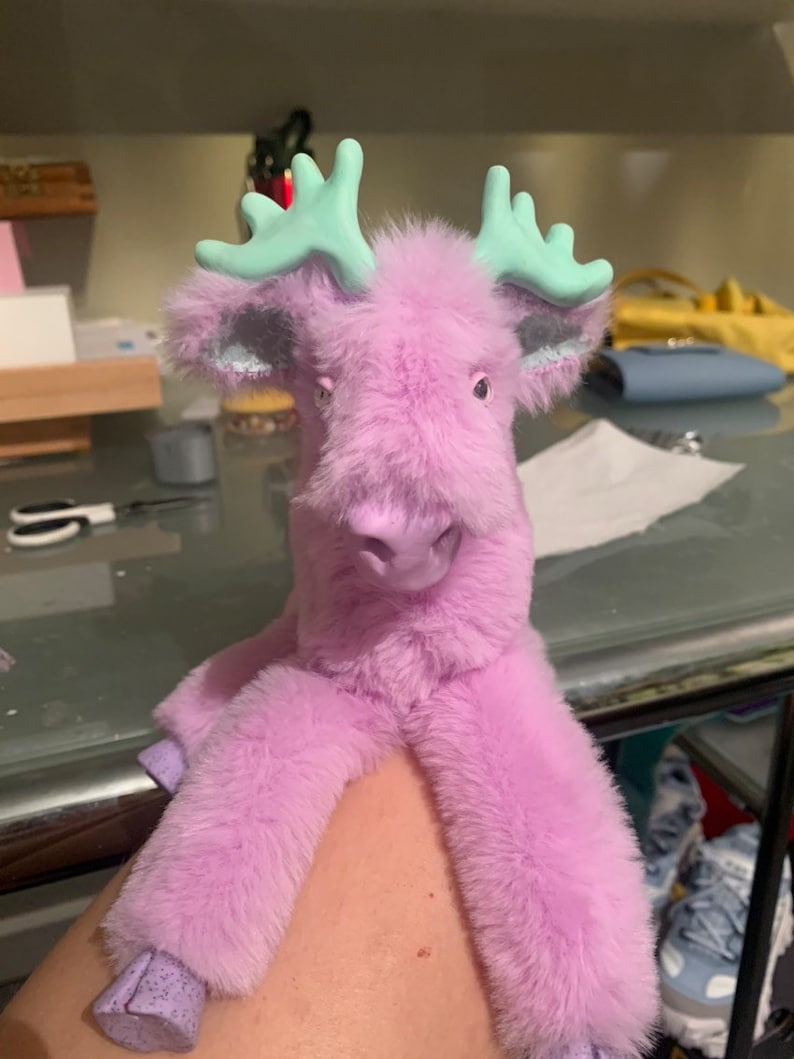 May include: A plush, lavender-colored moose toy with mint green antlers. The toy has a soft, furry texture and a friendly expression. The moose's hooves are a lighter shade of purple.