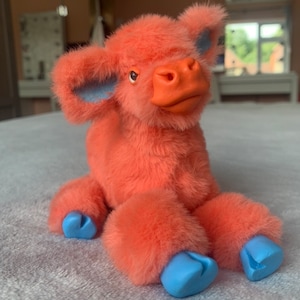 May include: A plush toy cow in shades of coral and blue. The cow has coral fur, a coral snout, and blue accents on the ears and hooves. The toy is sitting on a gray surface.