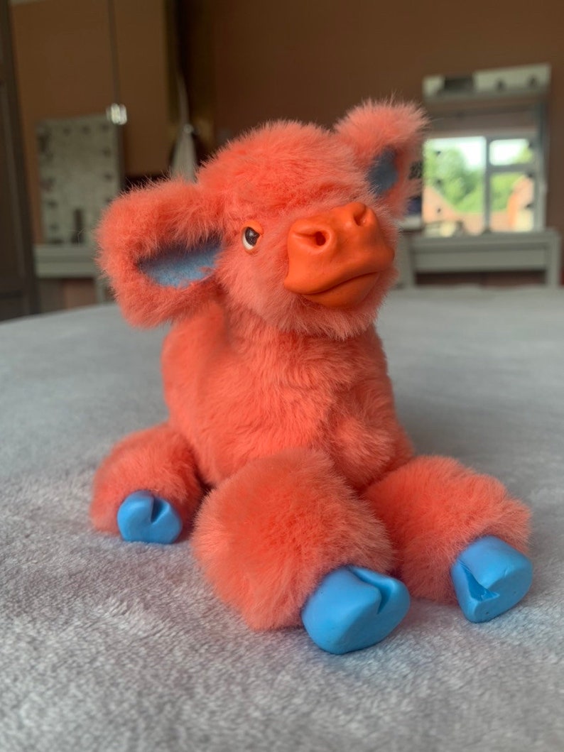 May include: A plush toy cow in shades of coral and blue. The cow has coral fur, a coral snout, and blue accents on the ears and hooves. The toy is sitting on a gray surface.