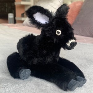 May include: A plush black deer toy with white-lined ears, a white muzzle, and a single eye visible. The toy is sitting on a soft, gray surface, with its legs extended. The deer has a fluffy, textured appearance.