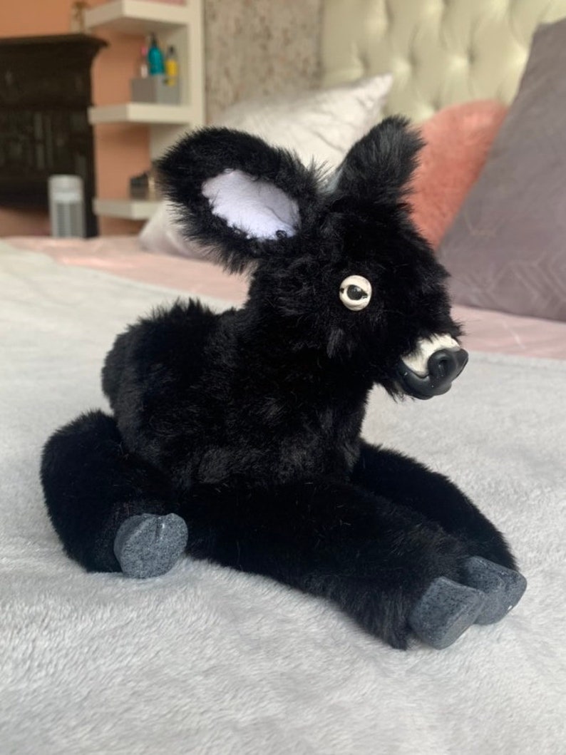 May include: A plush black deer toy with white-lined ears, a white muzzle, and a single eye visible. The toy is sitting on a soft, gray surface, with its legs extended. The deer has a fluffy, textured appearance.