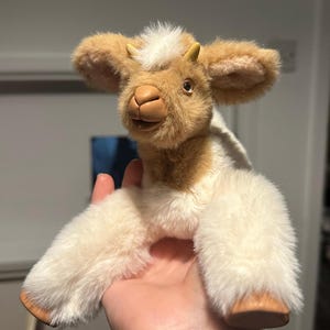 May include: A plush goat toy with a cream and brown color scheme. The goat has a soft, fluffy texture, small horns, and large ears. The toy is held in a hand, showcasing its size and detail. The goat's face has a friendly expression.