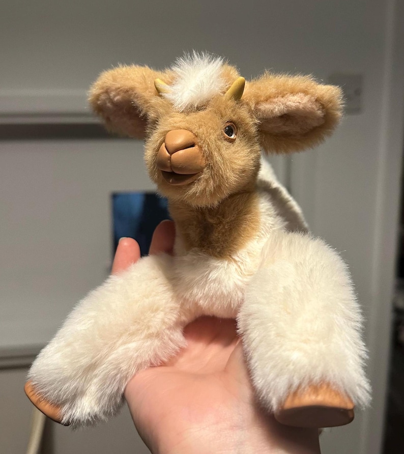 May include: A plush goat toy with a cream and brown color scheme. The goat has a soft, fluffy texture, small horns, and large ears. The toy is held in a hand, showcasing its size and detail. The goat's face has a friendly expression.