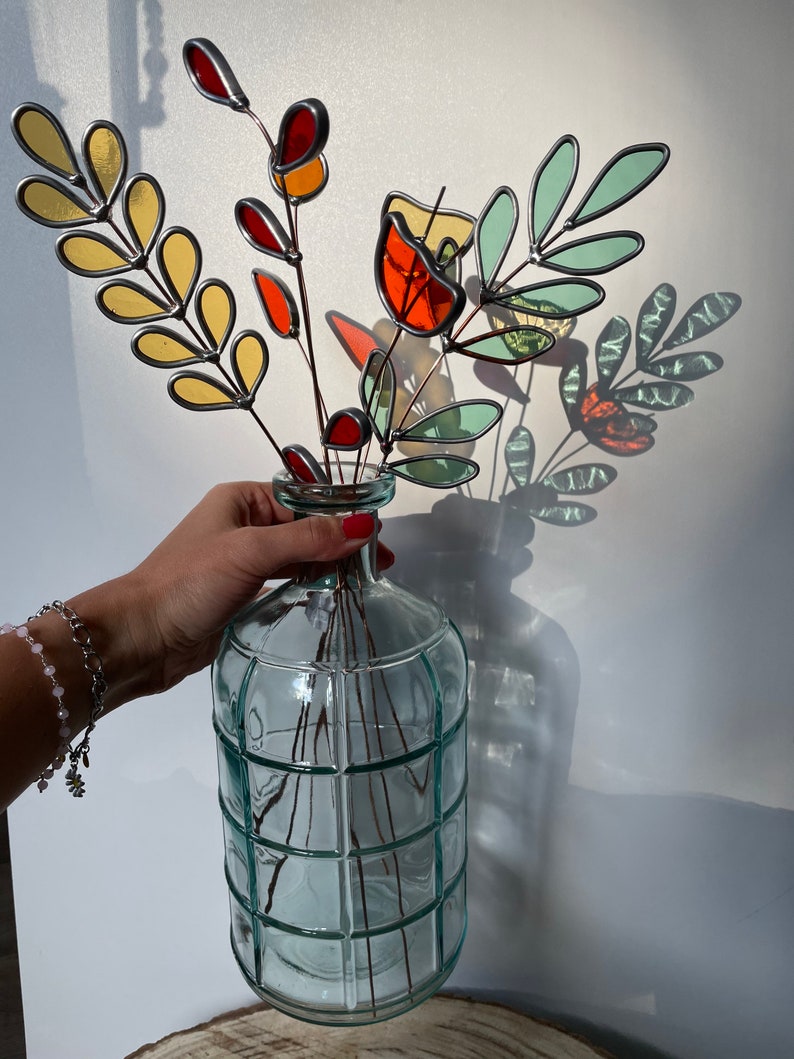 May include: A bouquet of stained glass flowers in shades of red, orange, yellow, and green, arranged in a clear glass vase with a grid pattern.