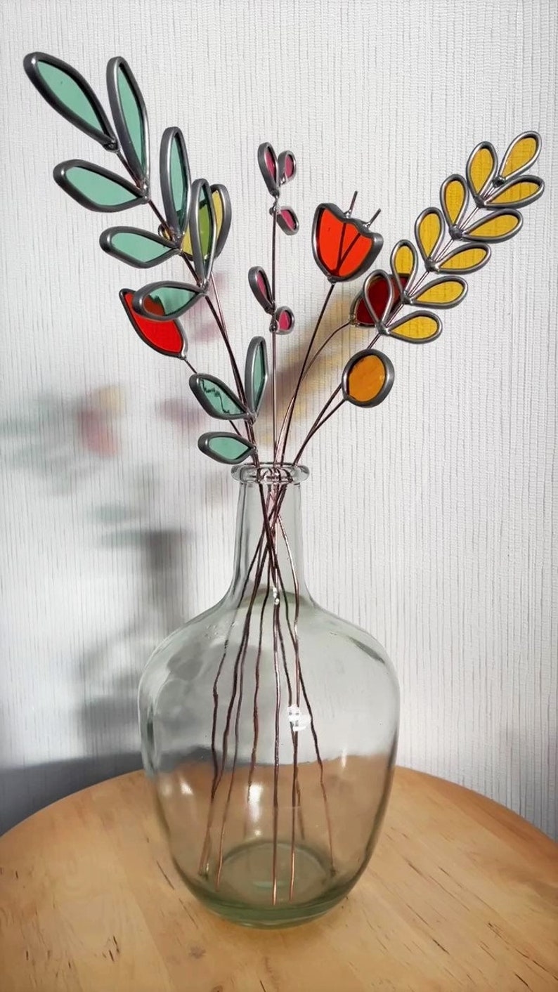 May include: A glass vase with a bouquet of colorful stained glass flowers. The flowers are made of different colors of glass, including red, orange, yellow, and green. The vase is clear glass and has a round shape.