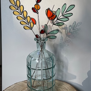 May include: A bouquet of stained glass flowers in a clear glass vase with a textured surface. The flowers are made of yellow, orange, red, and green glass.