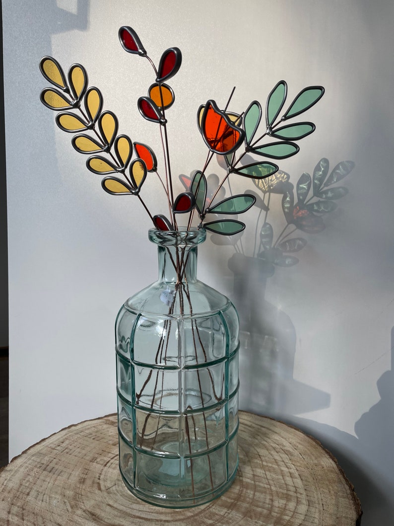 May include: A bouquet of stained glass flowers in a clear glass vase with a textured surface. The flowers are made of yellow, orange, red, and green glass.