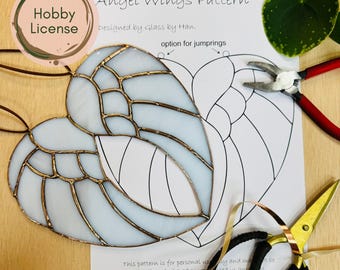 May include: A stained glass angel wing ornament with a light blue and gold design. The image includes a pattern sheet, pliers, and scissors. The text on the pattern reads "Angel Wings Pattern" and "Designed by Glass by Han."