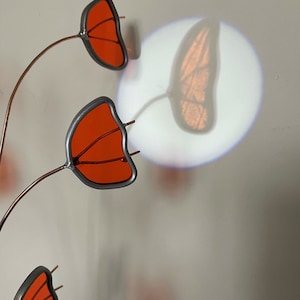May include: A metal sculpture with three orange glass butterfly wings attached to thin copper wires. The sculpture is casting a shadow of a butterfly on the wall behind it.