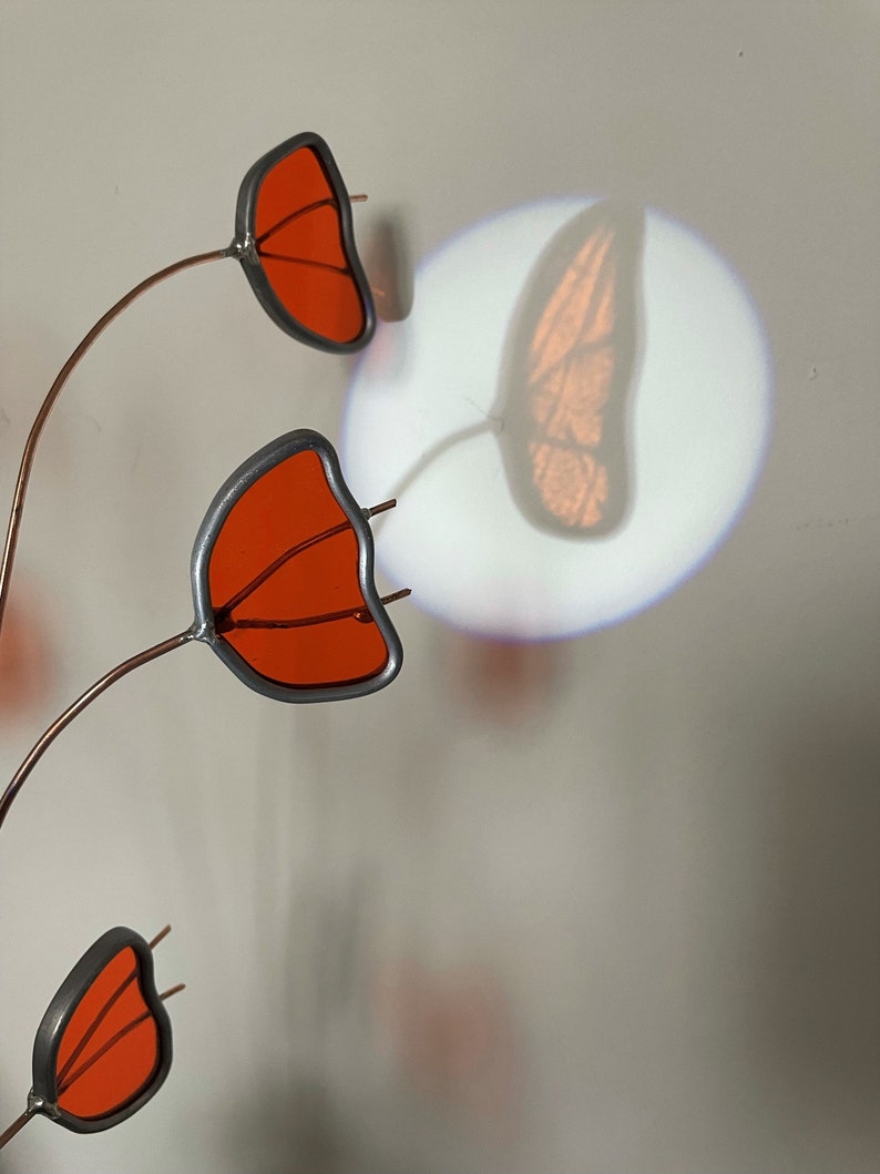 May include: A metal sculpture with three orange glass butterfly wings attached to thin copper wires. The sculpture is casting a shadow of a butterfly on the wall behind it.