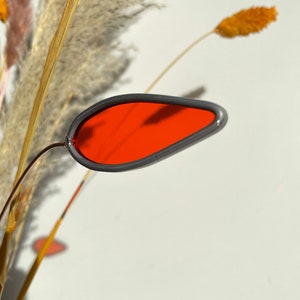 May include: A red glass teardrop-shaped ornament with a gray rim, hanging from a dried grass stem.