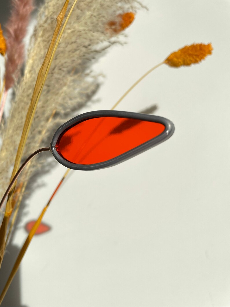May include: A red glass teardrop-shaped ornament with a gray rim, hanging from a dried grass stem.