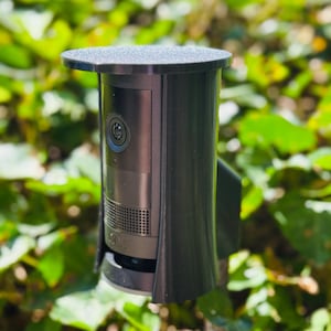 Puede incluir: Una cámara de seguridad negra con una cubierta protectora, montada en el exterior. La cámara tiene una lente y un altavoz, y está rodeada de follaje verde. La cubierta tiene una parte superior plana y texturizada.