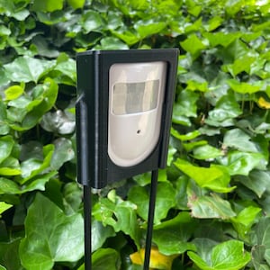 May include: A black plastic housing with a white motion sensor inside. The housing is mounted on two black metal stakes.