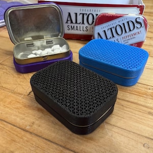 Puede incluir: Tres latas de mentas Altoids, una morada, una azul y una negra. Las latas están todas cerradas y tienen un diseño estampado en la parte superior.