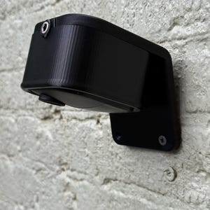 May include: A black security camera fixed to a white brick wall. The camera has a rectangular body with a curved front and a small lens. The mounting bracket is black and secured to the wall with screws.