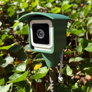 May include: A green weatherproof camera housing with a white camera inside. The camera is mounted on a metal pole and is surrounded by green foliage.