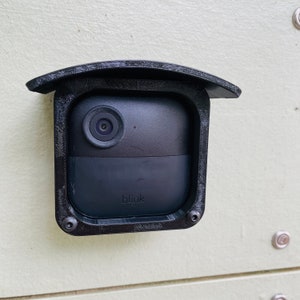 Puede incluir: Una cámara de seguridad Blink negra montada en una pared con una cubierta protectora negra. La cámara tiene una lente y el logotipo de Blink es visible.