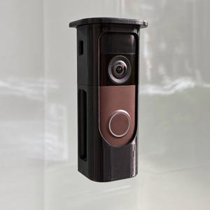 May include: A black rectangular security camera housing with a matte finish, featuring a central circular lens and a lower circular button. The device has a vertical orientation with a flat top edge and a smooth, structured exterior casing.