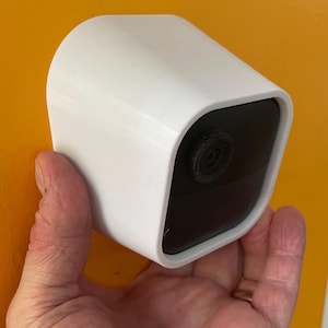 May include: A white security camera with a black lens is held in a hand. The camera has a rounded, rectangular shape and is designed for outdoor use.