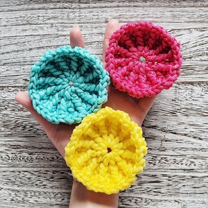 Puede incluir: Tres estropajos de ganchillo en diferentes colores: turquesa, rosa y amarillo. Cada estropajo tiene una forma circular con un agujero en el centro.