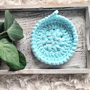 Peut inclure: Un panier rond en crochet bleu clair avec une poignée en boucle est posé sur un plateau en bois vieilli. Le plateau est gris clair avec des accents blancs. Des tiges de plantes vertes et feuillues sont à gauche, sur un fond blanc duveteux.
