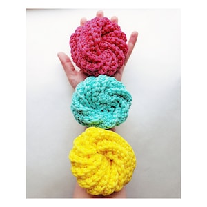 May include: Three crocheted hair scrunchies in a stack. The top scrunchie is pink, the middle scrunchie is teal, and the bottom scrunchie is yellow.