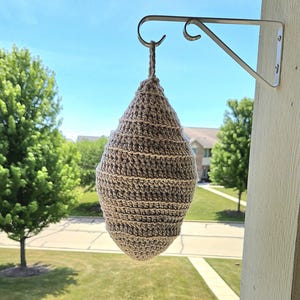 May include: A handmade, crochet birdhouse in a teardrop shape, suspended from a white metal bracket. The birdhouse is crafted from brown and tan yarn, with horizontal stripes. The background shows a green lawn, trees, and a blue sky.