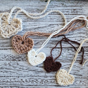 May include: Six crocheted hearts in various shades of brown and white. The hearts are strung together with twine and are hanging on a white wooden background.