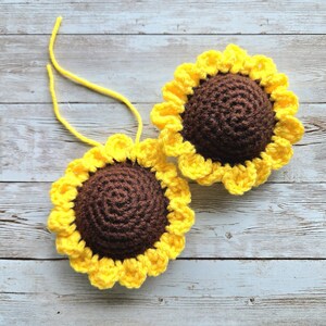 Op de afbeelding: Twee handgemaakte gehaakte zonnebloemornamenten. Elke zonnebloem heeft een bruin midden en gele bloemblaadjes. Aan elke zonnebloem is een geel touwtje bevestigd. De zonnebloemen worden gepresenteerd op een lichtblauwe en witte houten ondergrond.