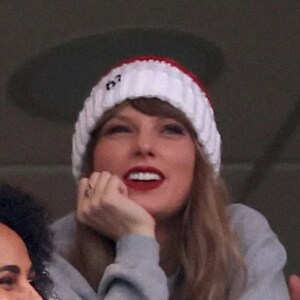 May include: A woman wearing a white and red knitted hat with the word "13" on it, a grey sweater, and long blonde hair. She is smiling and looking upwards.