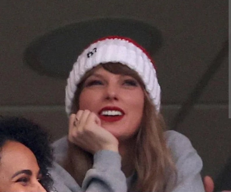 May include: A woman wearing a white and red knitted hat with the word "13" on it, a grey sweater, and long blonde hair. She is smiling and looking upwards.