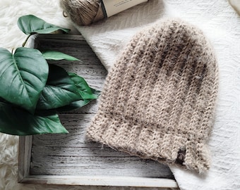 Op de afbeelding: Een beige gebreide beanie met een textuurpatroon. De beanie ligt op een wit houten dienblad met een groene plant op de achtergrond.