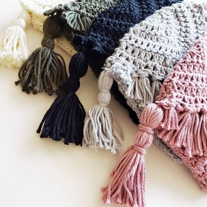 Puede incluir: Cinco gorros de crochet con borlas en varios colores, incluyendo blanco, verde, negro, gris y rosa.