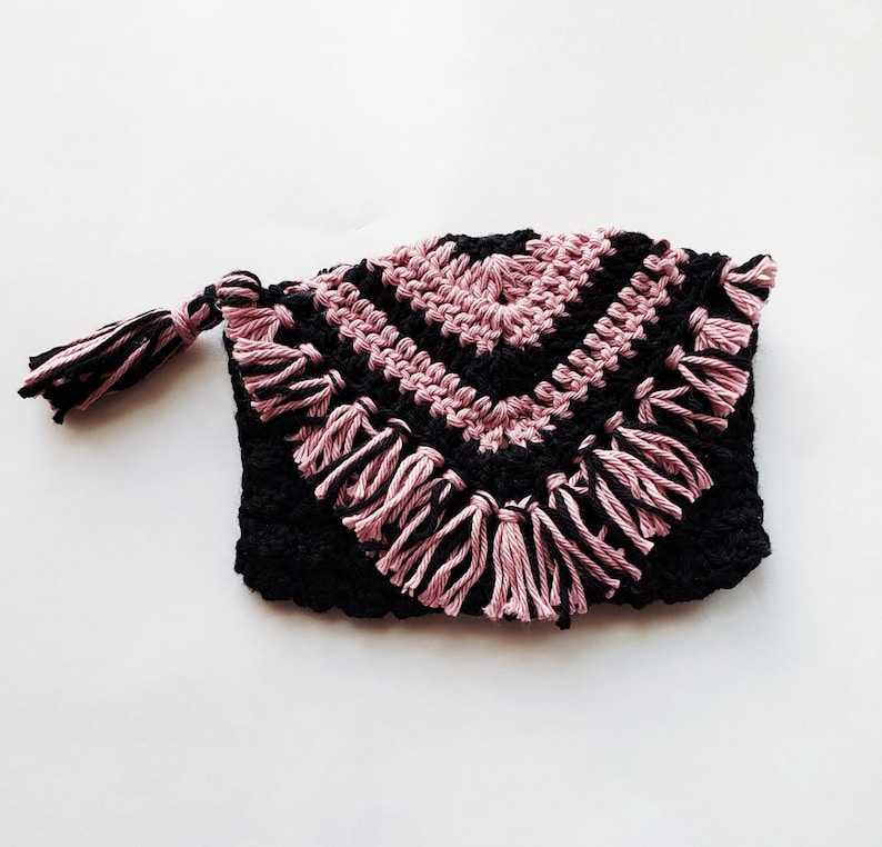 Puede incluir: Una bolsa de crochet negra y rosa con un borla. La bolsa tiene un patr&oacute;n geom&eacute;trico y un borde de flecos.