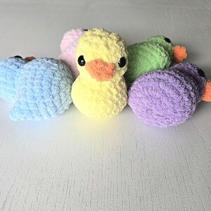 May include: A collection of six handmade, plush ducklings in various colors: yellow, blue, green, purple, and pink. Each duckling has black eyes and an orange beak. The ducklings are made of a soft, textured yarn.
