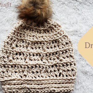 K&ouml;nnte beinhalten: Eine beige gestrickte M&uuml;tze mit einem braunen Kunstfellbommel. Die M&uuml;tze hat ein strukturiertes Strickmuster und hei&szlig;t "The Dreamsicle Beanie".