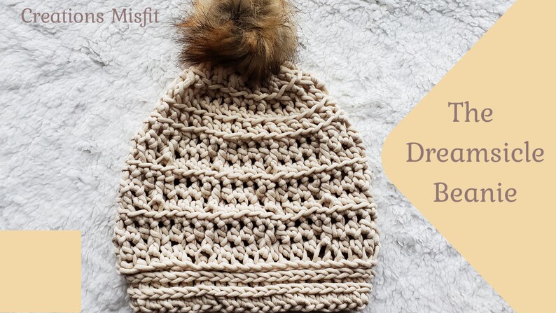 K&ouml;nnte beinhalten: Eine beige gestrickte M&uuml;tze mit einem braunen Kunstfellbommel. Die M&uuml;tze hat ein strukturiertes Strickmuster und hei&szlig;t "The Dreamsicle Beanie".