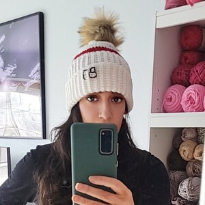 May include: White crocheted beanie with a red stripe and a faux fur pom pom. The beanie has the letters "FG" embroidered on the front.