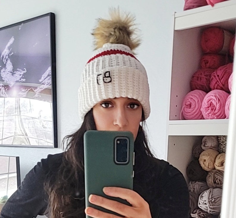 May include: White crocheted beanie with a red stripe and a faux fur pom pom. The beanie has the letters "FG" embroidered on the front.