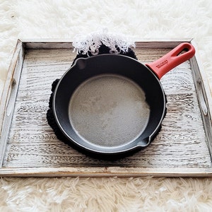Pode incluir: Uma pequena frigideira de ferro fundido preta com uma al&ccedil;a vermelha repousa sobre um descanso de panela preto de croch&ecirc;. A frigideira est&aacute; dentro de uma bandeja de madeira desgastada e branqueada. Uma borda branca com franjas &eacute; vis&iacute;vel.
