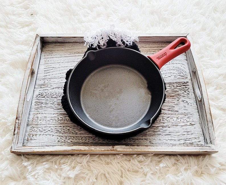 Pode incluir: Uma pequena frigideira de ferro fundido preta com uma al&ccedil;a vermelha repousa sobre um descanso de panela preto de croch&ecirc;. A frigideira est&aacute; dentro de uma bandeja de madeira desgastada e branqueada. Uma borda branca com franjas &eacute; vis&iacute;vel.