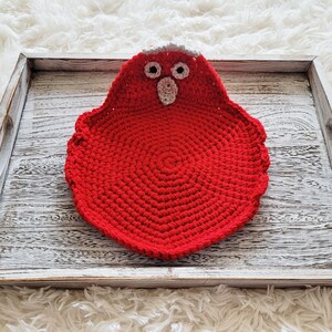 Pode incluir: Um descanso de panela feito &agrave; m&atilde;o, vermelho, em croch&ecirc; em forma de p&aacute;ssaro com detalhes cinza e branco. O descanso de panela &eacute; colocado em uma bandeja de madeira branqueada. O p&aacute;ssaro tem dois olhos pretos e um bico pequeno.