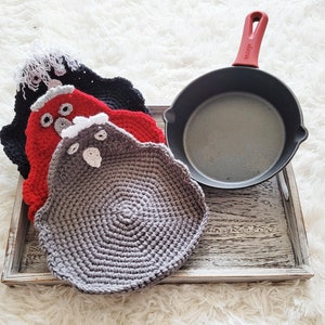 Pode incluir: Uma frigideira de ferro fundido com uma al&ccedil;a vermelha repousa numa bandeja de madeira junto a tr&ecirc;s pegadores de panela feitos &agrave; m&atilde;o em forma de galinha de croch&ecirc;. Os pegadores s&atilde;o pretos, vermelhos e cinzentos, com detalhes em branco e cinzento. A bandeja est&aacute; sobre uma superf&iacute;cie branca e fofa.