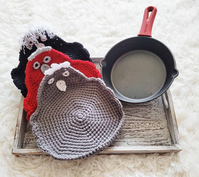 Pode incluir: Uma frigideira de ferro fundido com uma al&ccedil;a vermelha repousa numa bandeja de madeira junto a tr&ecirc;s pegadores de panela feitos &agrave; m&atilde;o em forma de galinha de croch&ecirc;. Os pegadores s&atilde;o pretos, vermelhos e cinzentos, com detalhes em branco e cinzento. A bandeja est&aacute; sobre uma superf&iacute;cie branca e fofa.