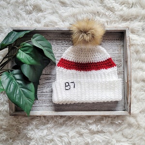 May include: White crocheted beanie with a red stripe and the number 87 embroidered on the front. The beanie has a fluffy pom pom on top.