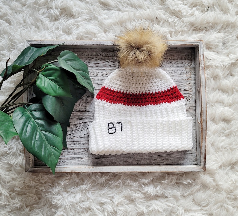 May include: White crocheted beanie with a red stripe and the number 87 embroidered on the front. The beanie has a fluffy pom pom on top.